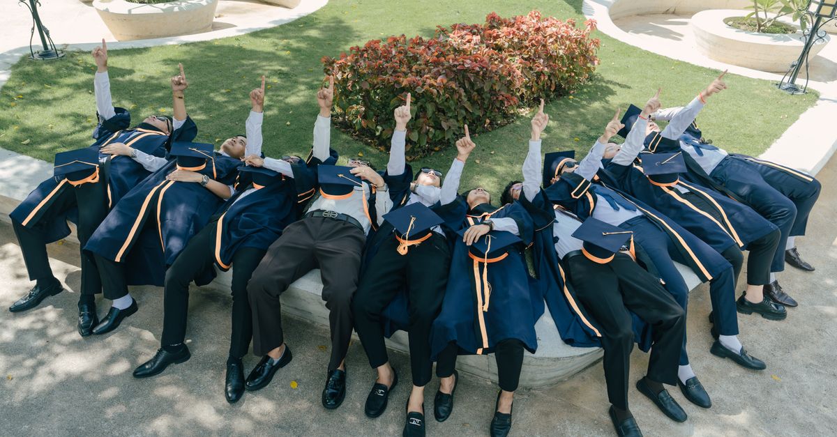 Graduates in caps and gowns celebrate by pointing skyward outdoors on a sunny day.